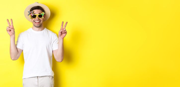 Concept of tourism and lifestyle. Happy man enjoying trip, wearing summer hat and sunglasses, posing with peace signs for photo, yellow background.