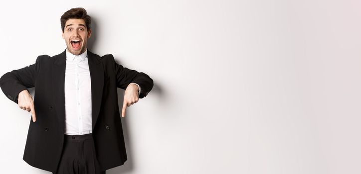 Portrait of super happy man showing christmas promo offer, pointing fingers down and smiling amazed, wearing formal party suit, standing over white background.