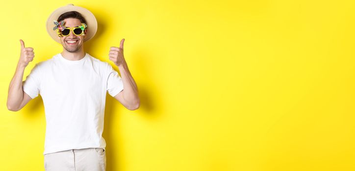 Concept of tourism and lifestyle. Image of smiling tourist showing thumbs-up, enjoying trip and recommending, wearing summer hat and sunglasses, yellow background.