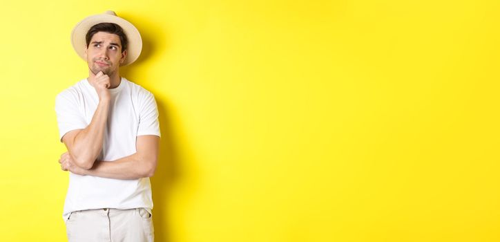 Concept of tourism and summer. Thoughtful man tourist pondering, looking at upper left corner and thinking, standing in straw hat and white t-shirt against yellow background.