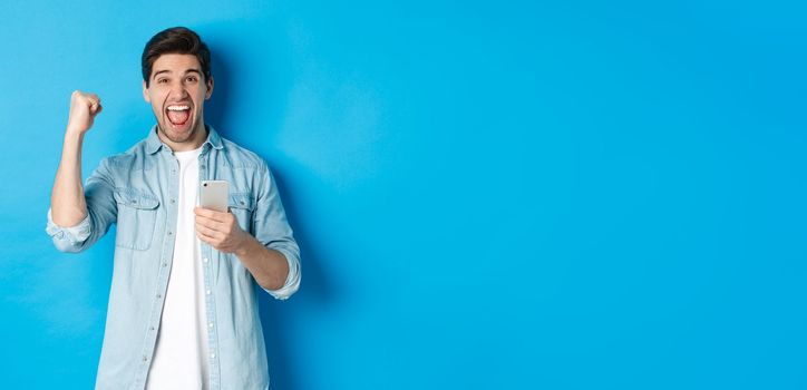 Concept of online shopping, applications and technology. Excited man shouting yes and making fist pump gesture after winning on smartphone, standing over blue background.