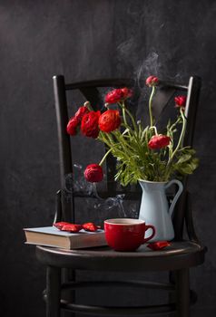 vintage still life on a black chair against a black background, red ranunculi in a blue jug, a red cup of tea with smoke, a blue book and candies in a red wrapper, dark photo, High quality photo