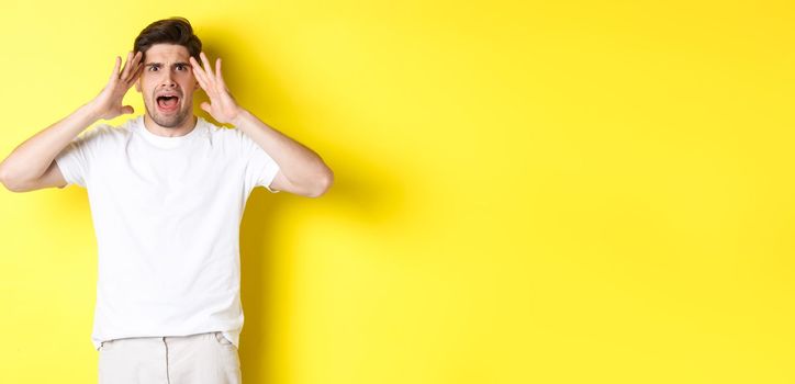 Frustrated guy looking alarmed, holding hands on head and feeling confused, panicking, standing over yellow background.