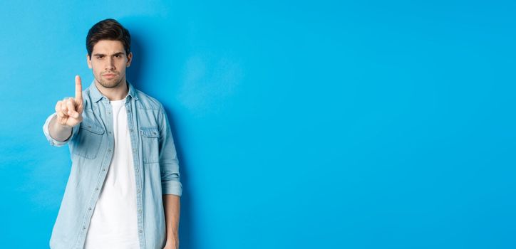 Angry 25 years old man shaking finger in disapproval sign, frowning disappointed, forbid something bad, telling no, standing against blue background.