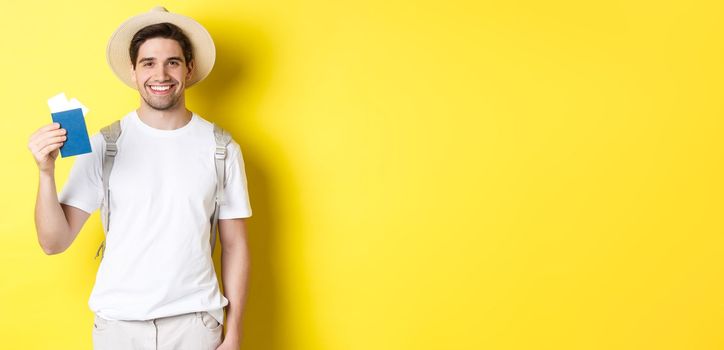 Tourism and vacation. Young smiling tourist showing passport with tickets, going on a trip, standing against yellow background.