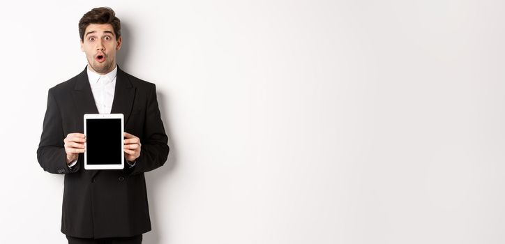 Image of surprised handsome man in black suit, showing digital tablet screen and looking amazed, standing against white background.