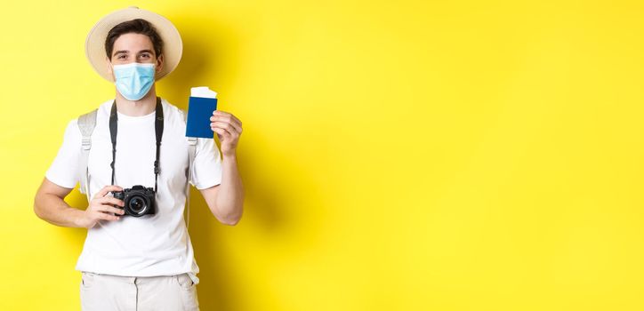 Concept of covid-19, travelling and quarantine. Happy man tourist with camera, showing passport and tickets for vacation, going on trip during pandemic, yellow background.