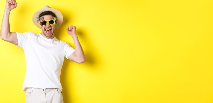 Concept of tourism and lifestyle. Happy lucky guy winning trip, rejoicing and wearing holiday outfit, summer hat and sunglasses, yellow background.