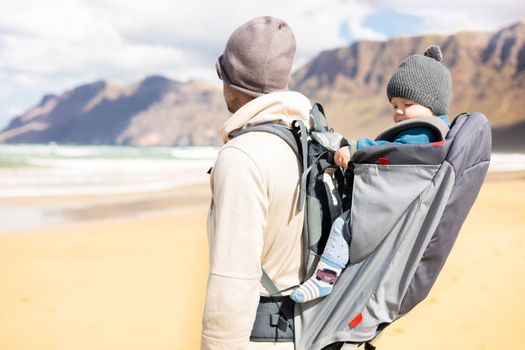 Young father carrying hisinf ant baby boy son in backpack on windy sandy Famara beach, Lanzarote island, Spain. Family travel and winter vacation concept