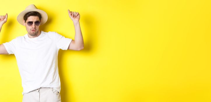 Tourism, travelling and holidays concept. Man tourist enjoying vacation, dancing in straw hat and sunglasses, posing against yellow background.