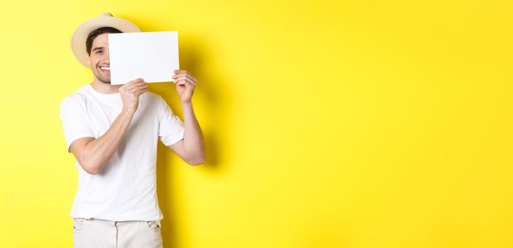 Happy guy on vacation showing blank piece of paper for your logo, holding sign near face and smiling, standing against yellow background.