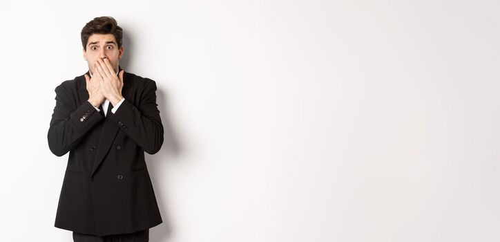 Scared man in formal suit, gasping and looking frightened at camera, standing against white background.