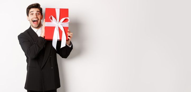 Image of happy good-looking man in black suit, holding box with christmas gift and smiling, standing against white background.