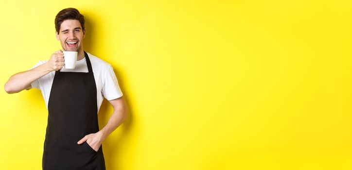 Handsome barista drinking cup of coffee and winking, inviting to cafe, standing over yellow background.