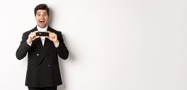 Portrait of amazed handsome man in black suit, showing credit card and recommending bank, standing over white background.