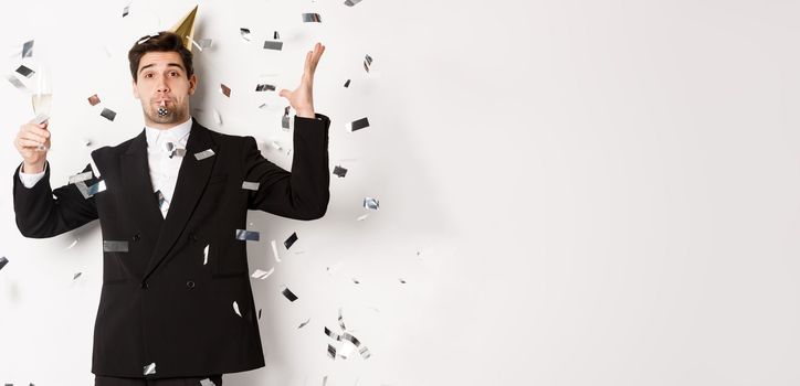 Handsome party guy in black suit having fun, celebrating new year, blowing whistle and drinking champagne while confetti falling, wishing happy holidays, standing against white background.