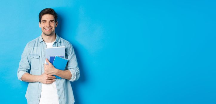 Young man holding notebooks and study material, smiling happy, standing over blue background.