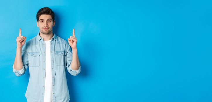 Image of calm handsome man showing you promo offer, pointing fingers up at copy space, standing against blue background.