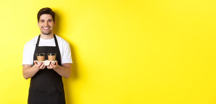 Handsome male barista serving takeaway coffee and smiling, bringing order, standing in black apron against yellow background.