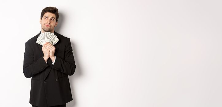 Image of handsome businessman in black suit, holding money and thinking about investment, making shopping plans, standing over white background.