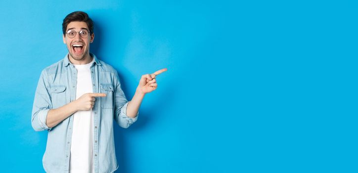 Surprised man in glasses pointing right at copy space, showing promo offer on blue background, standing over blue background.