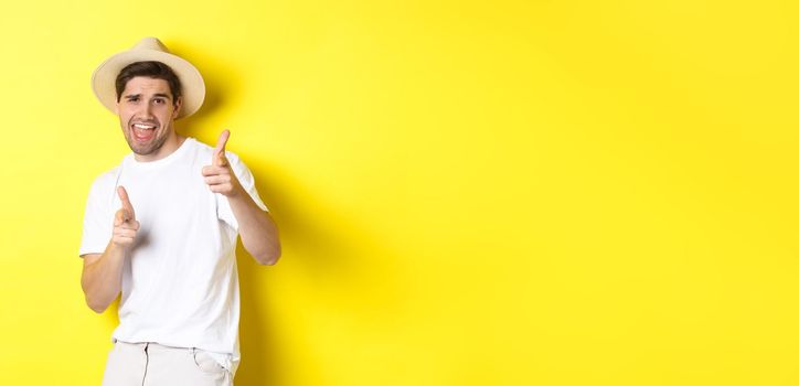 Concept of tourism and vacation. Happy cool guy pointing fingers at you, standing over yellow background.