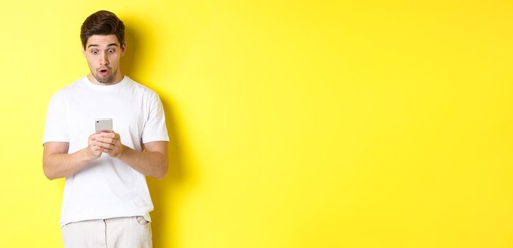 Man looking surprised in smartphone, reading message on cell phone, standing in white outfit against yellow background. Copy space