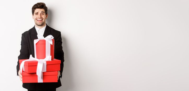 Image of handsome guy in black suit, holding gifts for christmas holidays, standing against white background.