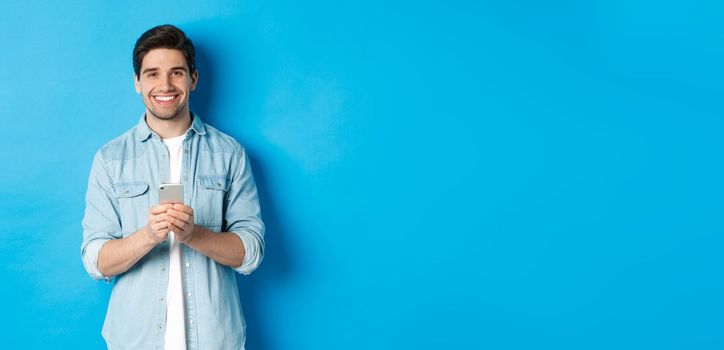 Handsome bearded man in casual outfit smiling at camera, checking smartphone, standing against blue background.