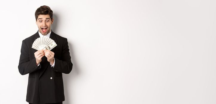 Image of handsome bearded guy in black suit, looking at money with excitement, standing over white background.