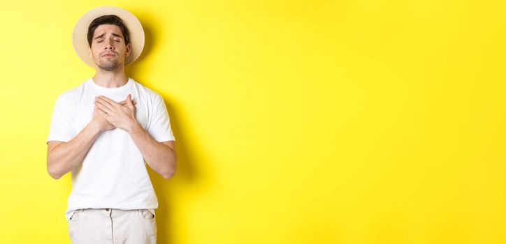 Concept of tourism and summer. Romantic man in straw hat looking nostalgic, close eyes and holding hands on heart, standing against yellow background.