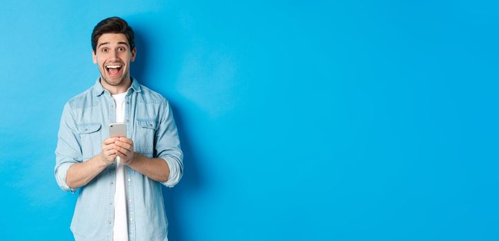 Surprised and happy man winning something online, holding smartphone and rejoicing, standing against blue background.