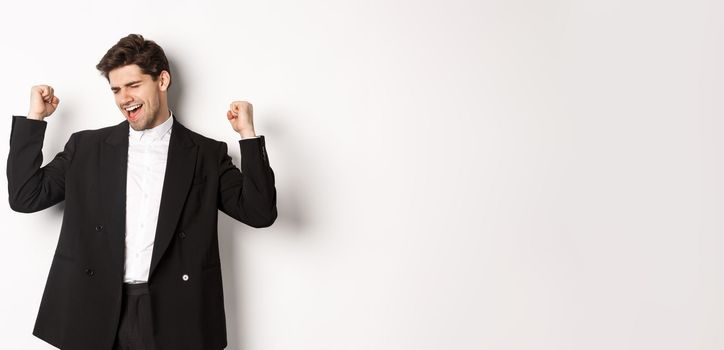 Image of successful and pleased handsome man in suit, rejoicing and making fist pump signs, dancing from happiness, standing over white background.