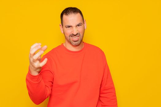 Middle age bearded man in red sweater gesturing claw with obvious angry, aggressive and thoughtful expression. Isolated on yellow background