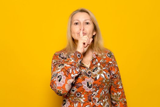 Pretty caucasian blonde woman in a patterned dress making the silence gesture with her index finger. She wants to create an aura of mystery, she hides a secret