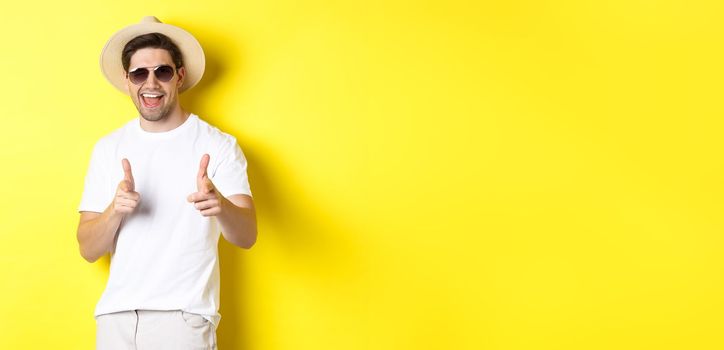 Confident and cheeky guy on vacation flirting with you, pointing finger at camera and winking, wearing summer hat with sunglasses, yellow background.