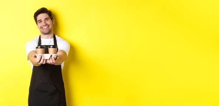 Friendly barista in black apron giving takeaway order, holding two cups of coffee and smiling, standing over yellow background.