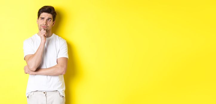 Portrait of young male model thinking, looking at upper left corner and making choice, standing near copy space, yellow background.