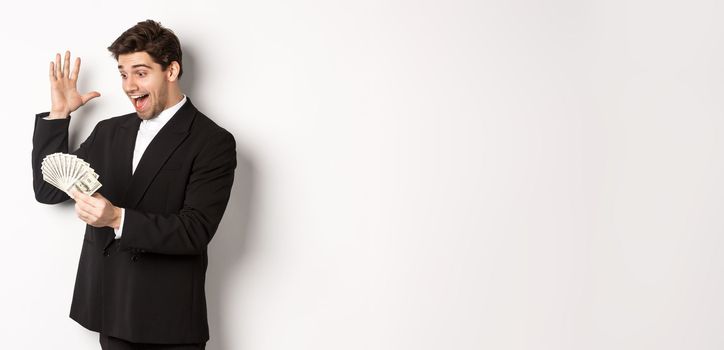 Image of successful businessman in black suit, looking at money and triumphing, earn cash, standing over white background.