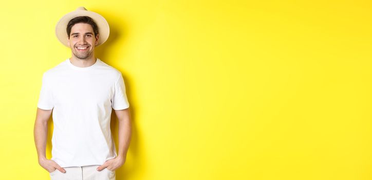 Young handsome guy tourist looking happy, wearing straw hat for travelling, standing against yellow background. Copy space