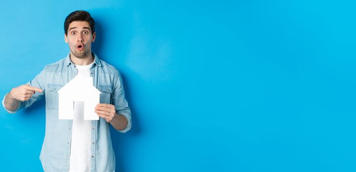 Insurance, mortgage and real estate concept. Surprised young man pointing at house card model and looking at camera, standing against blue background.