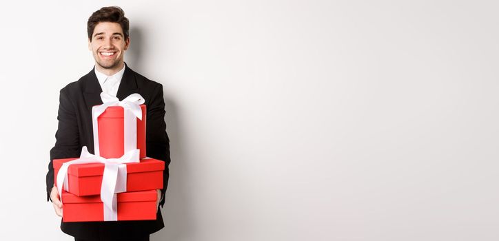 Portrait of handsome bearded man in trendy suit, holding gifts for new year and smiling, prepared presents, standing over white background.