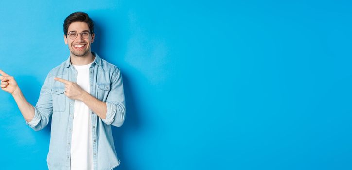 Young handsome man in glasses showing advertisement, smiling and pointing fingers left, making announcement, standing against blue background.