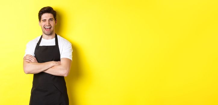 Handsome barista in black apron winking at you, wearing black apron uniform, standing over yellow background.