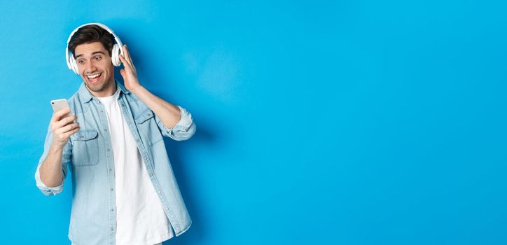 Happy man listening to music in headphones and reading message on smartphone, smiling excited, standing against blue background.