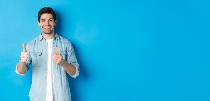 Concept of online shopping, applications and technology. Satisfied man in casual clothes smiling, showing thumbs up after using smartphone app, standing over blue background.