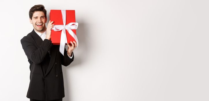 Concept of christmas holidays, celebration and lifestyle. Attractive man in black suit, holding new year gift and smiling, standing with a present over white background.