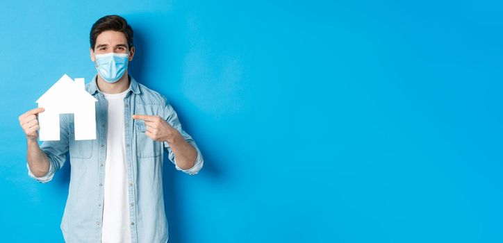 Concept of coronavirus, quarantine and social distancing. Young man searching aparment for rent, business loans, pointing at house model, wearing medical mask, blue background.