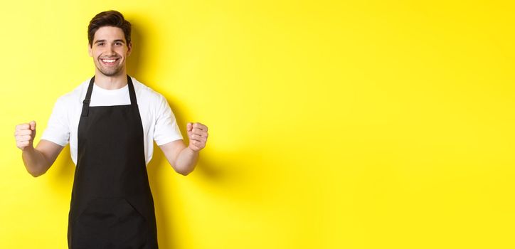 Friendly coffee shop waiter standing with raised hands, place for your sign or logo, standing over yellow background.