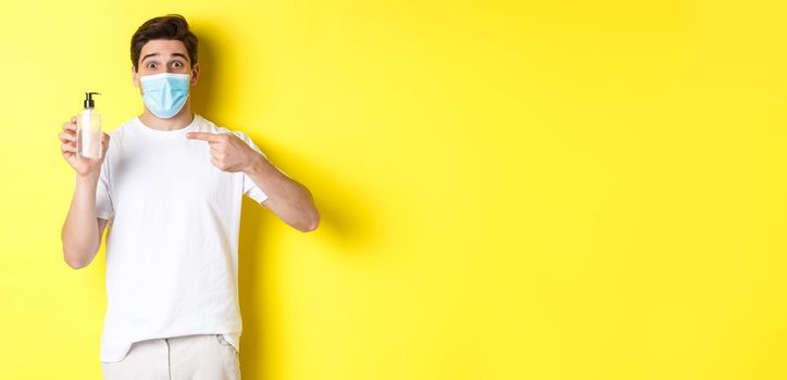 Concept of covid-19, quarantine and lifestyle. Excited guy in medical mask showing good hand sanitizer, pointing finger at antiseptic, standing over yellow background.